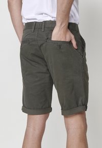 Vīrietis, kurš valkā olivu zaļas, sarullētas šortus, ar vienu roku aizmugures kabatā, redzams no vidukļa līdz ceļiem uz neitrāla fona.