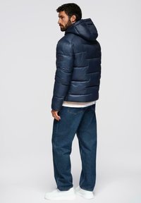 Giubbotto puffo blu navy con cappuccio, caratterizzato da sezioni trapuntate, tasche laterali e una texture liscia. Abbinato a jeans blu larghi e sneakers bianche.