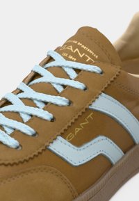 Καφέ δερμάτινα αθλητικά παπούτσια με ανοιχτό μπλε λεπτομέρειες και κορδόνια. Διαθέτουν οδοντωτές άκρες και χρυσό λογότυπο κοντά στη γλώσσα. Λαστιχένια σόλα.