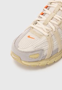 Gros plan d'une basket beige et gris clair avec tissu en mesh, lacets crème, un logo orange et une semelle texturée.