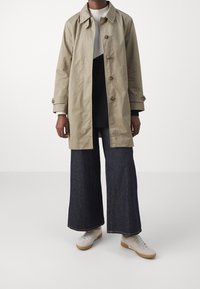 Trench-coat léger beige avec boutons, porté sur un pull sombre et un pantalon en denim large de couleur foncée, assorti à des chaussures blanches.