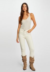 Un body sans manches beige clair scintillant avec un décolleté en V, assorti à un jean skinny crème et des bottines beige décontractées. Textures lisses tout au long.