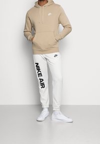 Felpa beige con cappuccio e tasca frontale; pantaloni della tuta bianchi con stampa nera "NIKE AIR"; sneakers bianche, sfondo uniforme.