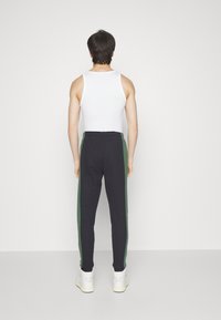 Herrsportkläder: svarta sweatpants med gröna sidostripe, resårmidja och åtsittande muddar, kombinerade med en vit linne och vita sneakers.