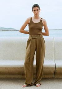 Bruine tanktop met een zijbanddetail, gecombineerd met hooggewaisted, loszittende beige broek. Witte slippers maken de outfit compleet.