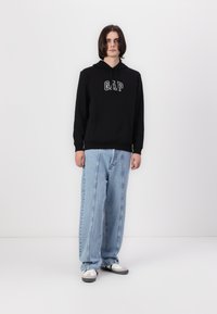 Sweat à capuche en coton noir avec le logo "GAP", associé à un pantalon en denim large bleu clair. Les chaussures présentent un schéma de couleurs noir et blanc.