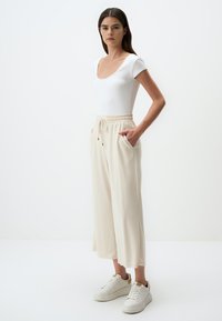 Witte geribde jumpsuit gecombineerd met crèmekleurige wijde sweatpants met een elastische tailleband en frontzakken; het model draagt witte sneakers.