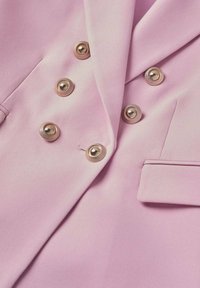 Blazer rose pastel avec six boutons métalliques ronds disposés en deux rangées verticales et une petite poche à rabat sur le côté droit.