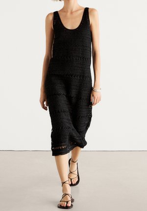 Femme portant une robe noire au crochet sans manches et des sandales noires lacées, debout sur un fond blanc uni.