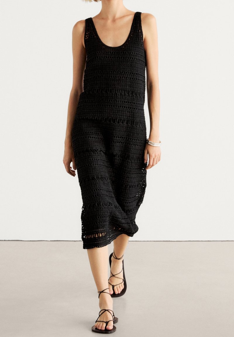 Femme portant une robe noire au crochet sans manches et des sandales noires lacées, debout sur un fond blanc uni.