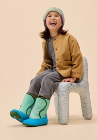 Grüne und blaue wasserdichte Stiefel mit einer strukturierten Oberfläche, getragen mit einer braunen Jacke über einem grauen Outfit, sitzend auf einem gepunkteten Stuhl.