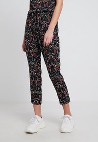 Kvinna i svarta, korta byxor med flerfärgad blommönster och vita, chunky sneakers, stående mot en ljus, enfärgad bakgrund.