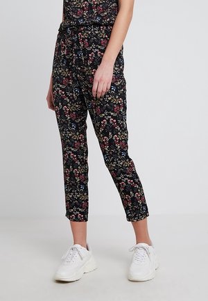 Femme portant un pantalon noir cropped avec un imprimé floral multicolore et des baskets blanches épaisses, se tenant devant un fond clair uni.