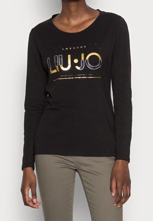 Person trägt ein schwarzes Langarmshirt mit dem gold- und silberfarbenen Schriftzug "LIU JO AWESOME" und hellgraue Hosen, vor weißem Hintergrund.