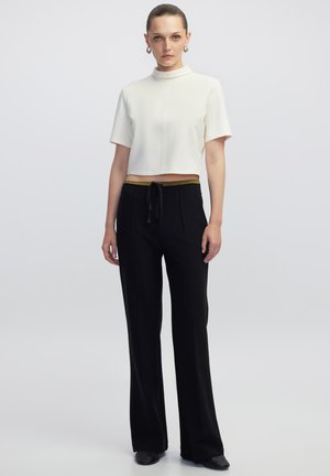 Witte cropped top met een hoge hals, gecombineerd met zwarte wijde broek met een olijfgroene tailleband en een strikkoord.