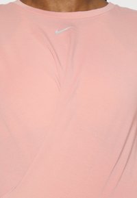 Vaaleanpunainen Nike-t-paita, jossa on sileä pinta, lyhyet hihat ja hopeinen logo rinnassa, sekä koristeellinen kierrekuvio.