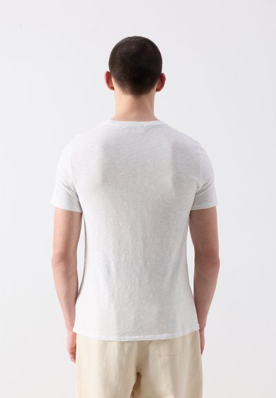 Homme aux cheveux courts et foncés portant une chemise blanche unie à manches courtes et un pantalon beige, vu de dos sur un fond blanc.