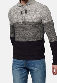 Man som bär en grå och svart stickad tröja med dragsko-krage och blå jeans, stående mot en enfärgad bakgrund.