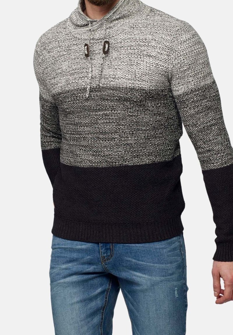 Man som bär en grå och svart stickad tröja med dragsko-krage och blå jeans, stående mot en enfärgad bakgrund.