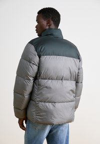 Siva puffer jakna s tamnozelenim gornjim delom, sa visokim ovratnikom i horizontalnim šavovima, izrađena od glatkog sintetičkog materijala.