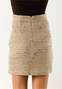 Minifalda beige de tweed de talle alto con sutiles motas marrones y cremallera visible en la parte trasera, combinada con un top negro acanalado.