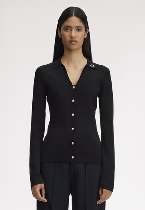 Femme aux longs cheveux foncés portant un cardigan noir cintré à boutons avec col et un pantalon noir, sur un fond clair uni.