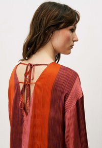 Blouse en soie à rayures verticales rouges et violettes, avec un décolleté en V et une fermeture nouée dans le dos. Texture douce et design fluide.