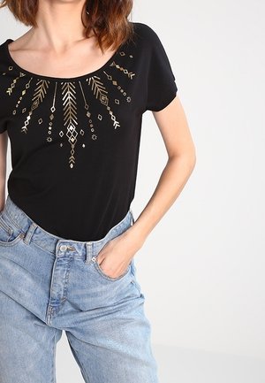 Nainen mustassa, kultaisin geometrisin kuvioin koristellussa pyöreäkauluksisessa paidassa ja vaaleansinisissä korkeavyötäröisissä farkuissa, käsi taskussa, selkeää taustaa vasten.