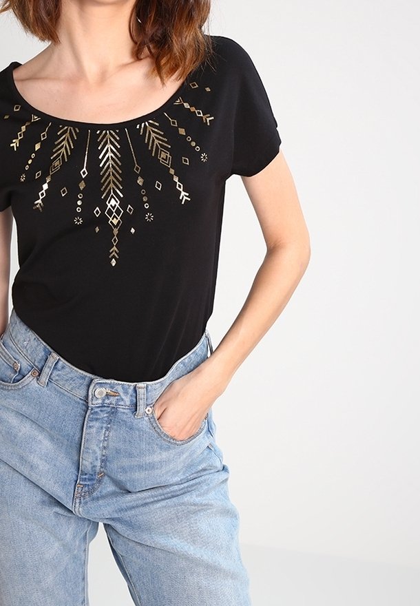 Femme portant un haut noir à col rond avec un motif géométrique doré et un jean taille haute bleu clair, main dans la poche, sur un fond uni.