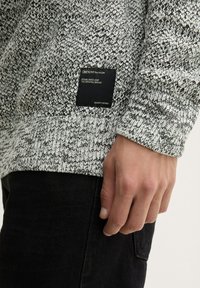 Pull en tricot dans des nuances de noir et blanc avec un motif texturé. Il comporte une étiquette de marque sur le côté et des poignets côtelés.