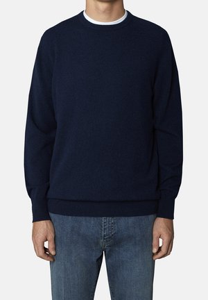 Mand iført en ensfarvet mørkeblå langærmet sweater over en hvid undertrøje, parret med lyseblå denimjeans, stående mod en hvid baggrund.