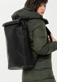Zaino a rotolo nero realizzato in materiale resistente, dotato di una tasca frontale con zip e spallacci regolabili, indossato su una giacca a vento verde.