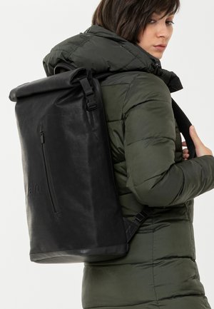 Schwarzer Rolltop-Rucksack aus strapazierfähigem Material, mit einer vorderen Reißverschlusstasche und verstellbaren Trägern, getragen über einer grünen Pufferjacke.