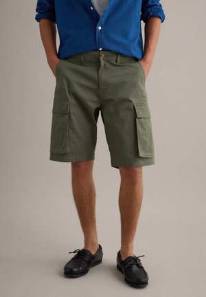 Uomo che indossa pantaloncini cargo verde oliva, una camicia blu con bottoni e maniche arrotolate, scarpe nere con lacci, in piedi con le mani nelle tasche.