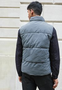 Grå polstret vest med høj krave og sorte ærmer. Stoffet virker blødt, med synlige syninger og en afslappet pasform.