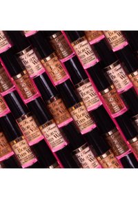 Des flacons de correcteur Too Faced Born This Way dans différentes teintes de peau, avec des accents dorés, de forme cylindrique et des bouchons noirs, présentés dans un agencement empilé.