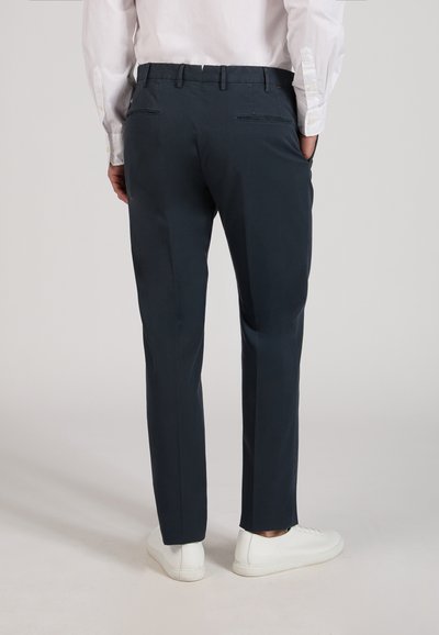 Man wearing dark navy trousers and white dress shirt, standing with hands in pockets, visible from back, wearing white sneakers.