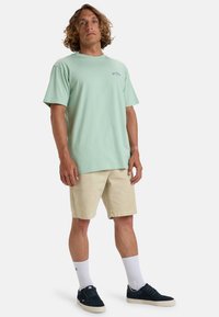 Mintgroen T-shirt met korte mouwen en een ronde hals, gecombineerd met beige shorts. Schoenen zijn marineblauw, met witte sokken zichtbaar boven de enkel.