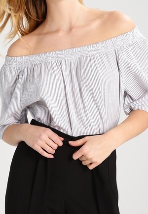 Kvinde iført en hvid og gråstribet skulderbar bluse og sort bukser med høj talje, hænder justerer linningen.