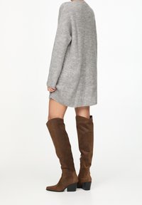 Bottes mi-mollet en daim marron avec un talon bloc, présentant une tige ajustée et un bout arrondi, assorties à une robe en maille grise.