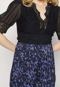 Top in pizzo nero con maniche a sbuffo trasparenti e scollatura a V, abbinato a una gonna con stampa leopardata blu e nera. Dettagli del tessuto con texture sono visibili.