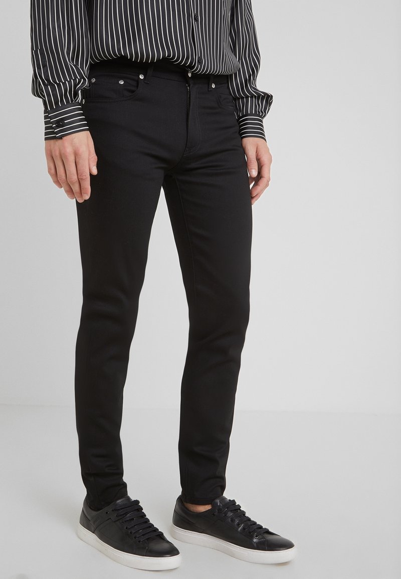 Jeans negros de corte slim, con cremallera, botones de metal y cinco bolsillos. Combinados con zapatillas negras y una camisa de rayas de manga larga.
