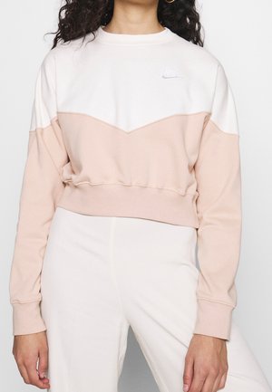 Kobieta ubrana w dwukolorową, beżowo-białą, krótką bluzę Nike z długimi rękawami oraz białe spodnie, stojąca na jednolitym tle.