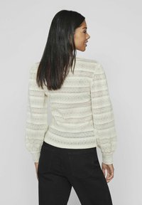 Gebreide trui in off-white met gestructureerde strepen, afgeronde schoudernaden en lange pofmouwen. Gepaarde met zwarte broek.