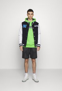 Schwarze und weiße Collegejacke mit blauer Schrift, neongrüner Kapuzenpullover, schwarze Shorts und weiße Sneaker mit silbernen Akzenten.