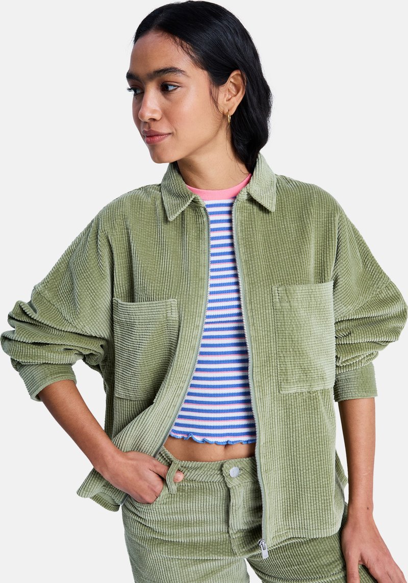 Πανωφόρι από βελουτέ ύφασμα σε ελιά πράσινο χρώμα, με υπερμεγέ θη εφαρμογή, δύο μπροστινές τσέπες και υφή, φορεμένο πάνω από ριγέ κορμάκι και παντελόνι στο ίδιο σχέδιο.
