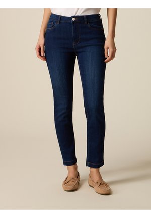 Kvinde iført mørkeblå slim fit jeans og beige mokassinsko, stående mod en ensartet lys baggrund.