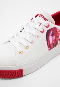 Baskets en toile blanche avec des accents de semelle rouges, des œillets multicolores, une doublure intérieure rouge et un motif floral rose-rouge sur le côté.