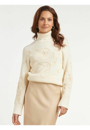 Vrouw met golvend bruin haar die een crèmekleurige coltrui draagt met getextureerde draaipatronen en een beige rok tegen een effen achtergrond.