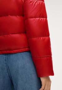 Czerwona kurtka puchowa z pikowanym wzorem, gładką, błyszczącą teksturą i dopasowanym rękawem. Nosi się ją na jasnoniebieskich dżinsach, eksponując elastyczny dół.